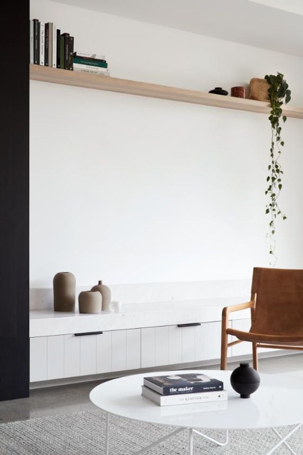 A calm, minimalist living room with a long white built-in sideboard and open pale timber display shelf against a white wall. Books, ceramics, woven storage and a trailing plant sit on the shelf, while a brown leather and timber armchair occupies the right side. A round white coffee table with stacked books and a black ceramic vase stands over a textured grey rug. A dark vertical architectural panel frames the left edge.