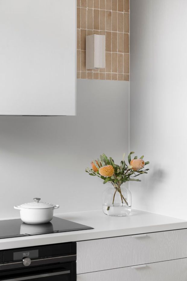 A close-up of a refined contemporary kitchen featuring pale grey cabinetry, a white benchtop, black built-in oven and induction cooktop, and a vertical backsplash of warm beige rectangular tiles. A white pot and glass vase with flowers sit on the benchtop. The space has clean lines, minimal detailing, and a soft neutral palette.