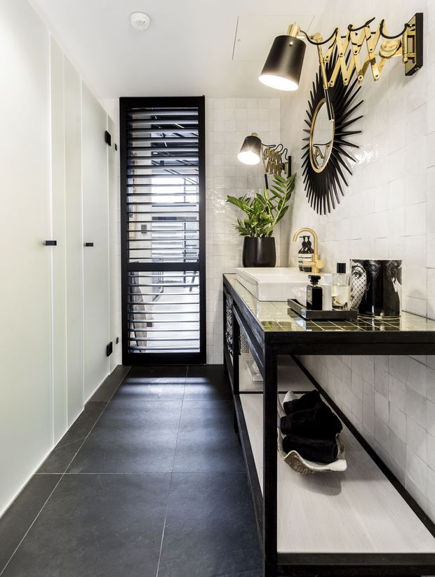 A narrow, contemporary bathroom with a long black vanity and white basin along a white tiled wall. The room features dark large-format floor tiles, white vertical wall panelling, a black-framed louvred door, brass-and-black wall lighting, and a striking sunburst mirror. A potted plant and neatly arranged toiletries add contrast to the monochrome and metallic palette.