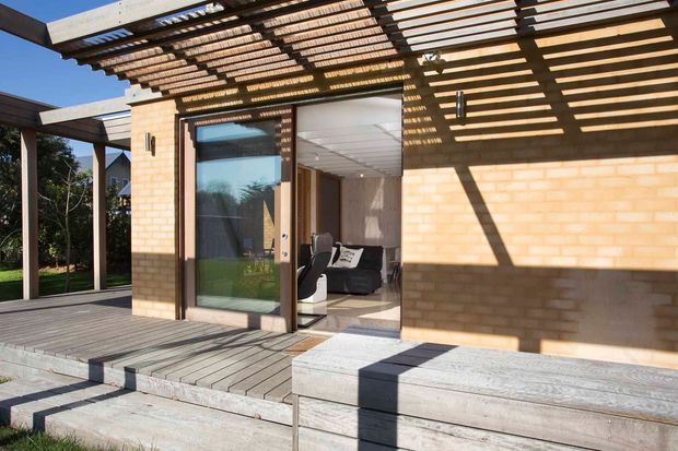 Exterior view of a contemporary brick-clad house opening onto a broad timber deck and stepped outdoor seating. A large glazed sliding door reveals part of a living room, while a timber pergola with closely spaced slatted beams casts strong linear shadows across the facade and deck.