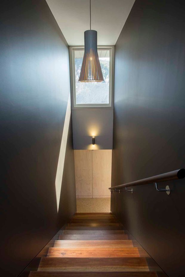 A narrow interior stairway descending through a dark, dramatic stairwell. Warm timber stair treads contrast with deep charcoal or dark grey walls, while a timber handrail and metal brackets run along the right side. A large pendant light hangs in front of a high window, with a small wall sconce illuminating the lower landing. Pale timber wall panelling and a terrazzo-like surface are visible at the bottom.