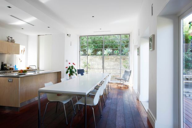 Bright open-plan kitchen and dining area with white walls, a long white dining table, timber kitchen cabinetry, stainless-steel appliances and a dark timber floor. Large glazed doors and windows open toward a leafy outdoor area, creating an airy connection to the exterior.