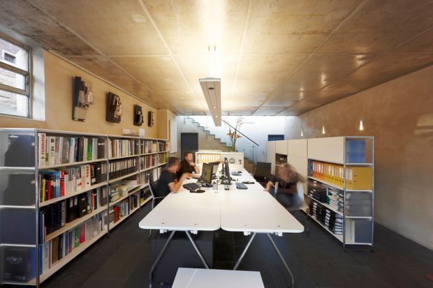 An open-plan office workspace in a long, narrow room, with shared white desks, office chairs, computers, extensive shelving and binders along both side walls, and a stairway leading to a brighter rear area. The space has a warm timber-panelled ceiling and wall surfaces, dark flooring, exposed shelving, and linear fluorescent lighting. The image has a contemporary, functional studio-office character.