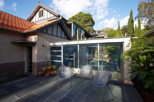 A sunny courtyard terrace adjoining a character residential house, with a glazed modern extension and outdoor seating. The original building features red terracotta tiled pitched roofs, rendered walls, dark brick basework, and decorative timber gable detailing, while the extension has extensive floor-to-ceiling glazing and a slim flat roof. Dark rectangular paving forms the terrace, surrounded by established garden planting.