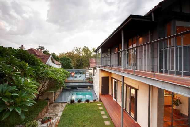Exterior view into a compact landscaped courtyard framed by a two-storey contemporary residential building. The home has dark roof planes, timber-clad balcony and deck surfaces, slender metal balustrades, large glazed doors and windows, and warm interior lighting. A small swimming pool, lawn, stepping stones, garden planting, and neighbouring pitched-roof dwellings are visible.