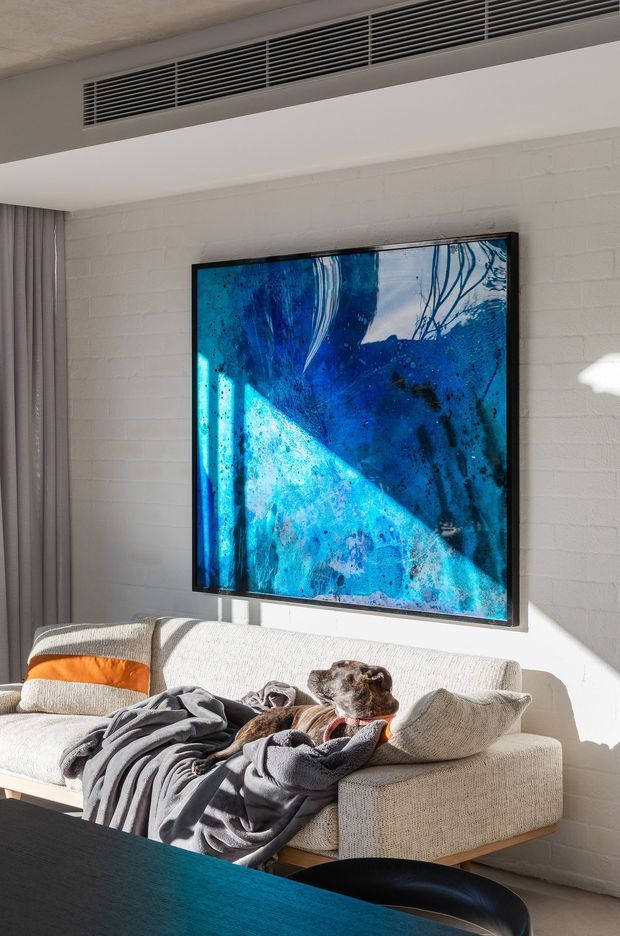 Bright contemporary living room with a white-painted brick feature wall, large blue abstract artwork, neutral textured sectional sofa, grey curtains, and strong sunlight creating geometric shadows. A dog is resting on the sofa, with a dark table visible in the foreground and a linear air-conditioning grille near the ceiling.
