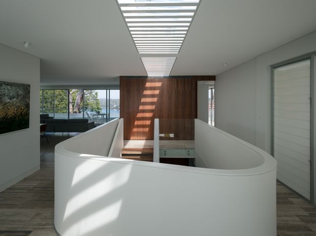 Contemporary interior view of an open stairway or landing, defined by a sculptural curved white balustrade around a stair void. A linear skylight or clerestory above casts strong striped sunlight onto a dark timber feature wall and timber flooring. Glazing at the rear overlooks greenery and water, while a living area is visible beyond.