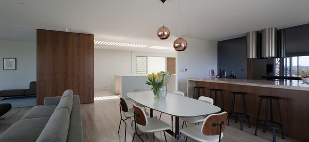 Open-plan contemporary kitchen, dining and living area with a light neutral palette. The space features a long white dining table with mixed upholstered and timber chairs, a grey sofa, timber flooring, dark cabinetry, a timber kitchen island with bar seating, and two reflective pendant lights. A partial timber divider separates the living zone from the dining area, while large glazing at the right brings in natural light and views.