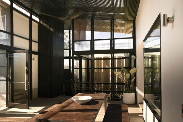 A contemporary interior courtyard or entry space with a long timber dining table, a shallow ceramic bowl, and a potted plant. Black steel-framed glazing and doors create a series of transparent partitions, while a ribbed dark ceiling and dark vertical wall cladding add texture. Strong sunlight enters through the glazing, and a large wall mirror reflects the space.