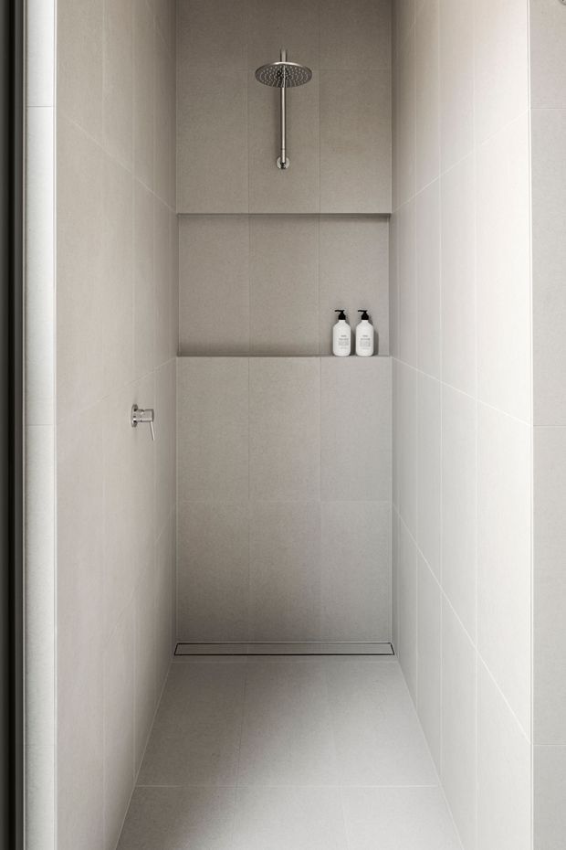 A narrow, minimalist walk-in shower clad entirely in light grey tiles, with a ceiling-mounted round rainfall showerhead, recessed wall niche containing two bottles, wall control, and linear floor drain. The space is clean, monochromatic, and softly lit.