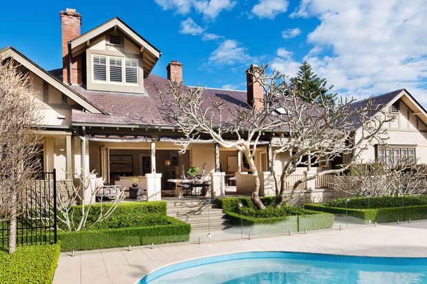 Exterior view of a large traditional residence with multiple gabled roof sections, prominent brick chimneys, timber detailing, dormer windows, and a covered veranda. Formal clipped hedges and a swimming pool occupy the foreground under a bright blue sky.