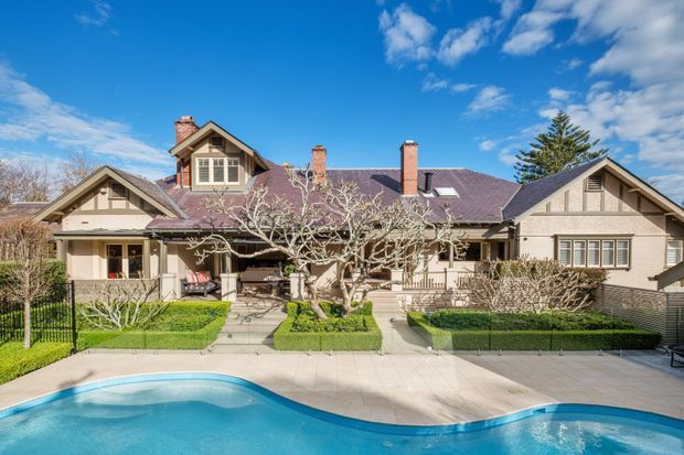 Front exterior view of a large single-storey residence with multiple gabled roof forms, brick chimneys, pale rendered and timber-trimmed façades, landscaped formal gardens, and a swimming pool in the foreground. The architecture has strong Craftsman and Californian bungalow influences, with broad eaves, exposed gable detailing, grouped windows, and a covered verandah.