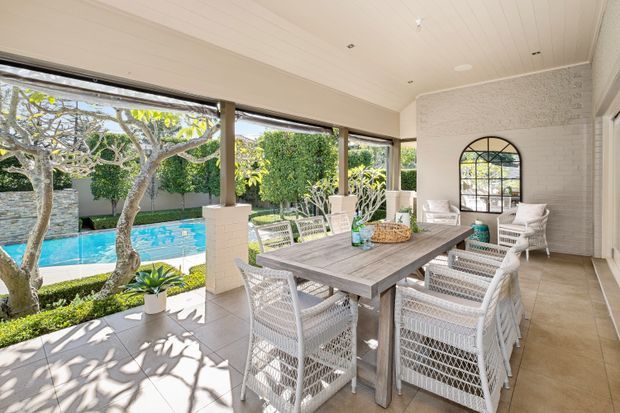 Bright covered outdoor dining terrace beside a swimming pool, with a large timber dining table surrounded by woven white chairs. The space features tiled flooring, white-painted brick columns and walls, a pale painted timber-lined ceiling, and broad open glazing overlooking landscaped gardens and the pool. An arched black-framed window adds a distinctive feature.