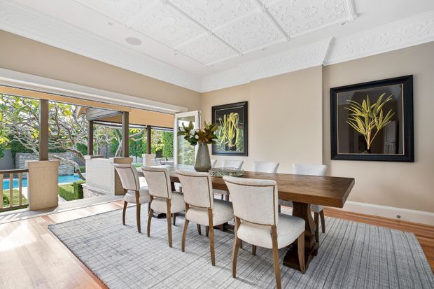 Bright, refined dining room with a long timber dining table and upholstered dining chairs on a large grey rug. The room features ornate white pressed-metal ceiling panels and decorative cornices, warm neutral walls, framed botanical artwork, timber flooring, and expansive glazed doors opening to a covered outdoor area and pool beyond.