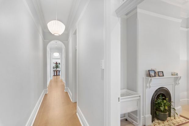 Bright, narrow traditional hallway with pale timber flooring, white painted walls, detailed architraves and cornices, and a series of arched openings leading toward a small console table. A white decorative fireplace with framed photographs and a potted plant occupies the adjoining room, while built-in cabinetry is visible beside the hallway. The space has a refined, airy, heritage-inspired character.