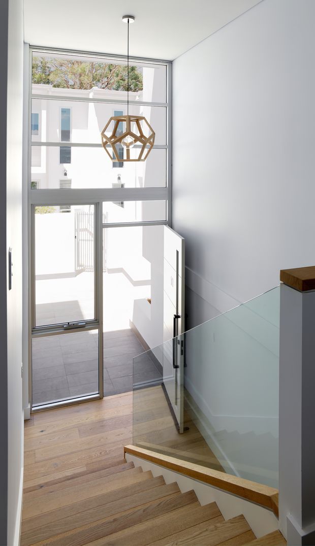 Bright contemporary stairway descending toward a glazed entry door, with wide floor-to-ceiling windows, pale timber stair treads and flooring, a clear glass balustrade with timber handrail, white walls, and a geometric timber pendant light. The glazing overlooks a light-filled courtyard or adjacent building.