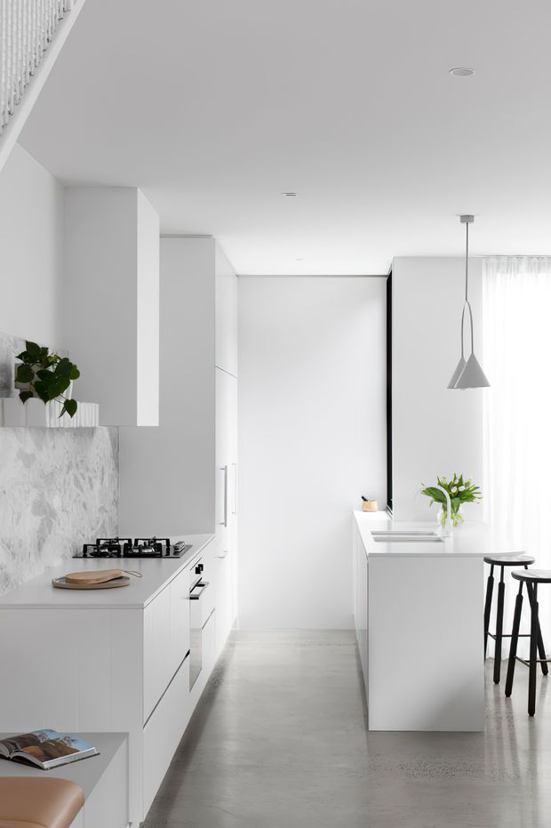 Bright, narrow contemporary kitchen with a minimalist white fit-out, full-height cabinetry, a pale grey marble-look splashback, white island bench, black bar stools, and a simple geometric pendant light. A large window with sheer curtains provides abundant natural light, while polished grey concrete flooring adds contrast and texture.