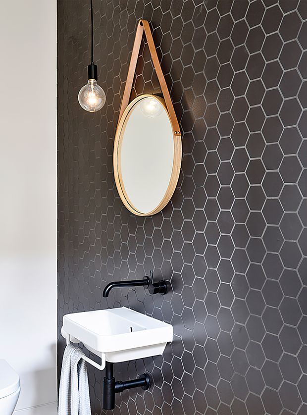 A compact bathroom or powder room with a wall-mounted white basin, black wall-mounted tapware and exposed plumbing, a dark grey hexagonal tile feature wall, and a small oval mirror suspended by a timber strap. A globe pendant light hangs beside the mirror, while part of a white toilet is visible at the left edge. The design has a strong monochrome palette with warm timber and soft-textured accents.