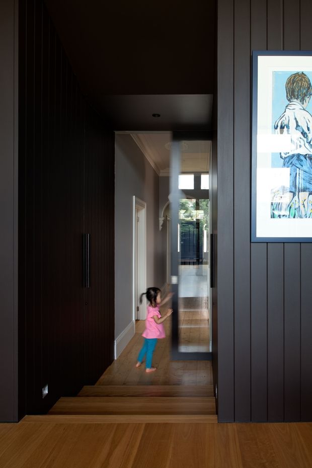 A dark, refined residential hallway viewed from a timber-floored landing, with a short set of timber stairs descending into the corridor. The hall features deep charcoal vertical wall panelling and concealed cabinetry with long black handles on the left, a framed artwork on the right, a glazed internal door or partition, and glimpses of bright rooms beyond. Warm timber flooring contrasts with the moody dark walls, while a child provides a sense of scale.