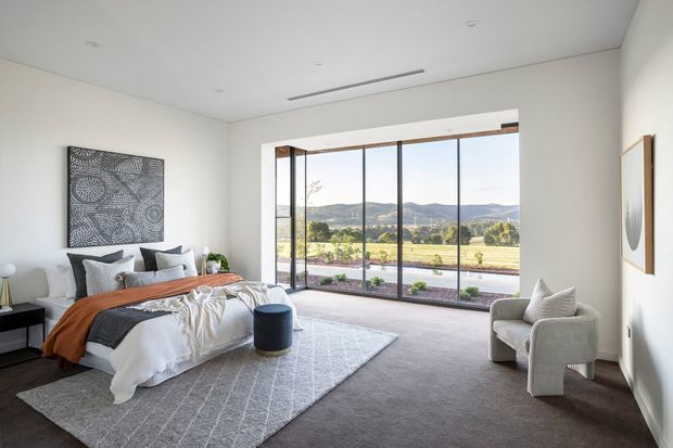 A bright, spacious contemporary bedroom with a neatly styled bed, neutral upholstered armchair, dark bedside furniture, a large textured area rug, and minimalist wall art. Floor-to-ceiling glazed sliding doors open the room to an expansive landscape view, while recessed ceiling lights and a linear slot diffuser maintain a clean, understated interior.
