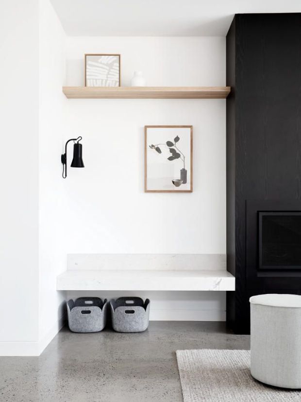 A minimalist contemporary living room corner featuring a white built-in desk or bench beneath a slim timber shelf, framed botanical artwork, a black wall sconce, and a tall dark built-in fireplace wall. The space has a pale polished concrete floor, soft neutral furnishings, and a clean monochromatic palette with warm wood accents.