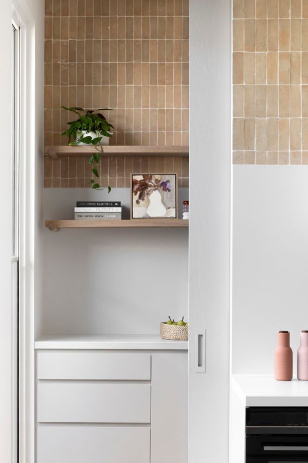 A bright, compact built-in kitchen storage area with white cabinetry, drawers, open shelving, and a warm beige tiled feature wall. Timber-toned shelves display a trailing plant, books, and framed artwork, while part of a kitchen benchtop and dark appliance are visible at right. The space has a calm, refined contemporary feel.