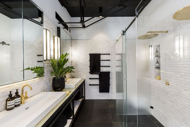 A contemporary bathroom with a long double vanity, white integrated basins, black open-frame cabinetry, and brass-toned tapware. White brick-pattern wall tiles contrast with large dark floor tiles and a black exposed ceiling. A frameless glass shower enclosure occupies the right side, with brass shower fittings and recessed wall niches. Large mirrors, vertical feature lighting, black towel rails, and indoor plants complete the space.