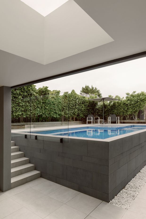 A modern covered outdoor pool area viewed from a tiled patio. The raised rectangular swimming pool has blue water, dark grey block-clad retaining walls, clear glass balustrades, and integrated tiled steps. A large white geometric roof or soffit shelters the foreground, while hedging, fencing, outdoor seating, and an umbrella are visible beyond the pool.
