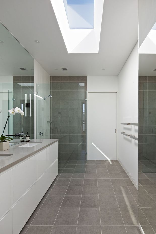 Bright contemporary bathroom with a long white double vanity, integrated basins, large wall mirror, glass-enclosed shower, grey tiled walls and floor, recessed ceiling lights, and a large skylight. White built-in cabinetry and open towel shelving create a clean, minimal appearance.