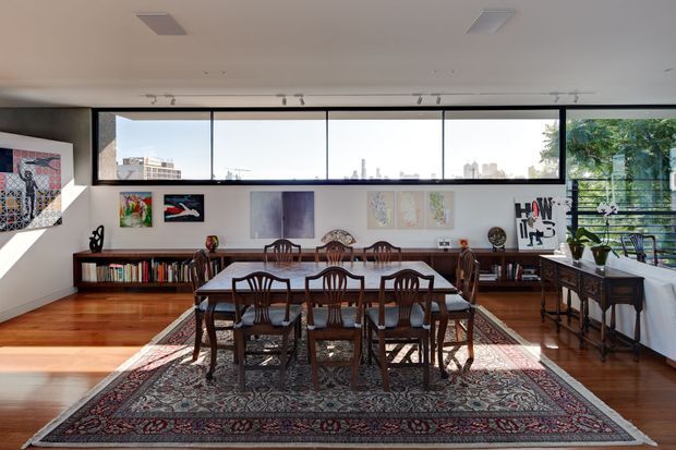 A bright, art-filled dining room with a long dark timber dining table and matching carved-back chairs arranged over a large patterned oriental rug. Built-in low bookshelves line the rear wall beneath a series of artworks, while a timber sideboard and decorative objects sit to the right. Full-width windows provide views toward a distant city skyline, with indoor plants and exterior greenery visible through the glazing. The room has polished timber flooring, white walls and ceiling, exposed picture rails or curtain tracks, and a contemporary open-plan feel.