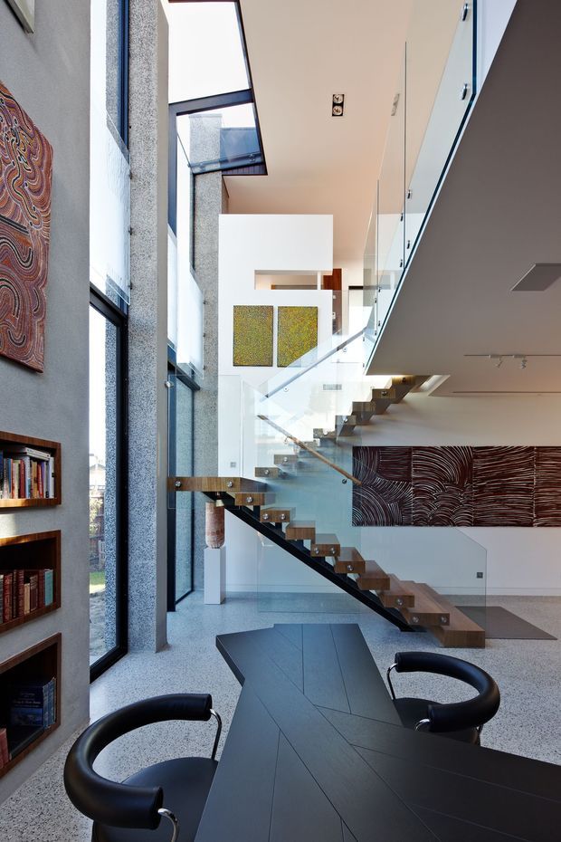 A tall, light-filled contemporary interior centred on a floating timber staircase with clear glass balustrades and a dark structural stringer. Full-height glazing and a double-height void create strong vertical openness. The lower level contains a dark dining table with black curved chairs, built-in timber bookshelves, polished aggregate-style flooring, and large abstract artworks in earthy tones. White walls and ceilings contrast with dark joinery and stair elements.