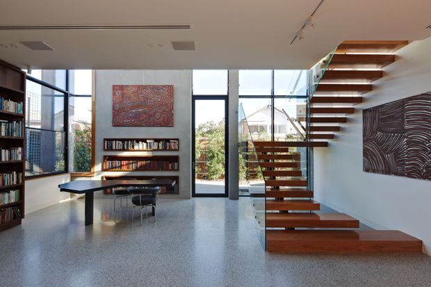 Bright contemporary open-plan interior featuring a sculptural floating timber staircase with glass balustrades, polished terrazzo-style flooring, full-height glazing, extensive built-in bookshelves, a compact dining/work table, and colourful wall art. The space has a refined minimalist character with strong natural light and a double-height stair volume.