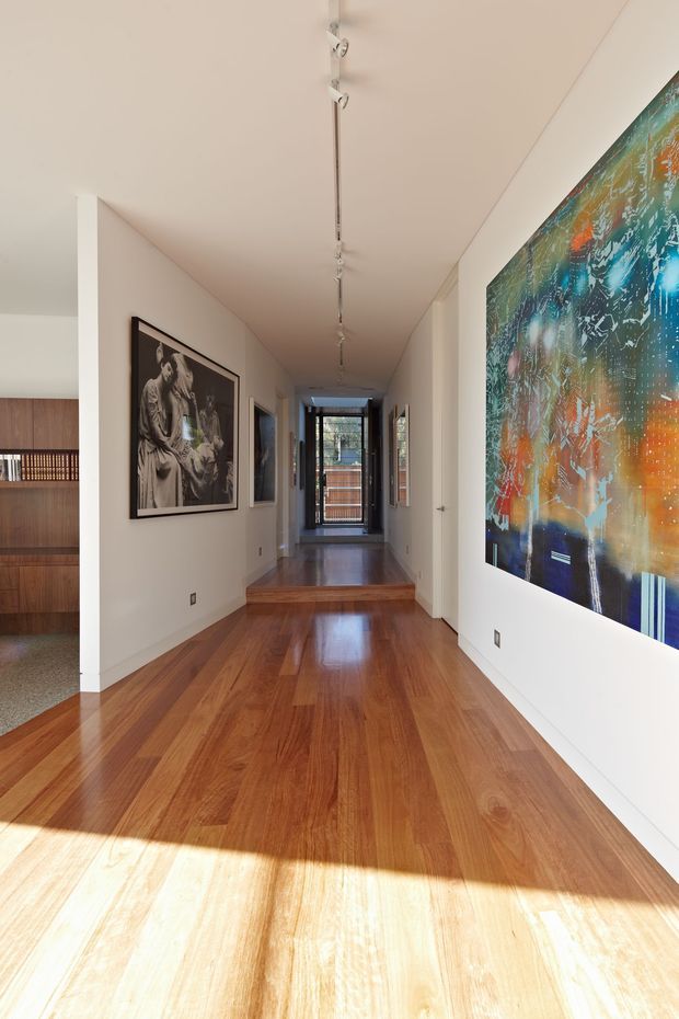 A bright, elongated residential hallway or gallery corridor with polished warm timber flooring, white walls and ceiling, recessed doorways, framed artwork, and a linear track light running along the ceiling. The corridor leads toward glazed doors or windows at the far end, with a timber-lined room partially visible through an opening on the left. Strong sunlight creates a bright foreground reflection on the floor.