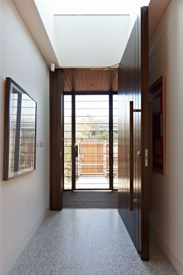A narrow, contemporary entry hall leading to a glazed rear doorway and small outdoor landing. The hall has a speckled terrazzo floor, smooth white walls, a large skylight, dark timber vertical-panel joinery and a timber-lined ceiling at the far end. Full-height black-framed glazing brings in daylight, while a framed artwork is visible on the left wall and partially behind the open timber door on the right.