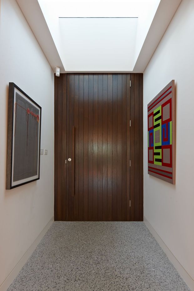 A narrow, bright entry hall focused on a tall dark timber pivot or hinged door with vertical board detailing. White walls and a large rectangular ceiling skylight create an airy, gallery-like space. The floor is finished in speckled terrazzo, with framed abstract artwork displayed on both side walls.