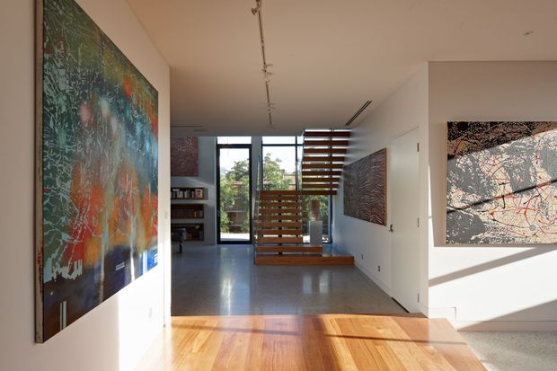 A bright contemporary interior hallway and living area with polished concrete flooring, warm timber floorboards in the foreground, white walls, large abstract artworks, and a floating timber-slat stair or screen leading toward a glazed rear door. Built-in shelving and concealed linear ceiling lighting add a refined architectural character.
