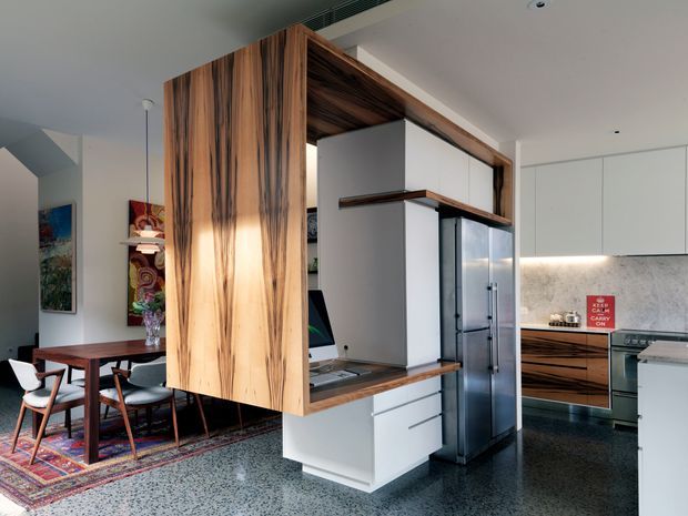 Open-plan contemporary kitchen with a large timber-lined island and partition feature dividing it from a dining area. White cabinetry, dark timber accents, stainless-steel appliances, marble-look splashback, and a polished terrazzo-style floor create a refined, warm modern interior. A dining room is visible to the left, with artwork and patterned rug adding colour.