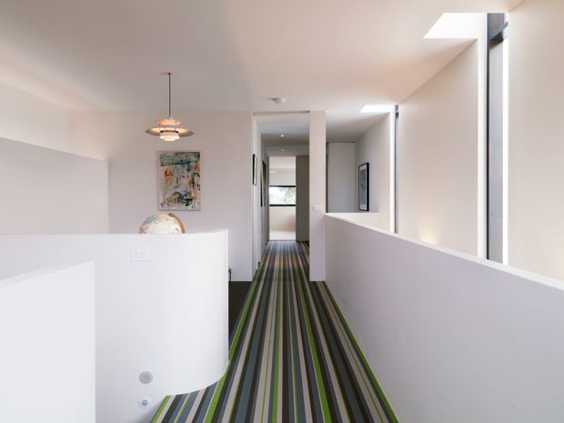 Bright contemporary interior hallway or landing with white walls, half-height balustrade walls, narrow vertical windows, and a vivid striped carpet runner in green, grey, black, and beige. A sculptural pendant light hangs near an abstract artwork, while a partially visible curved white built-in element occupies the foreground.