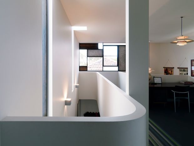 Interior view of a contemporary multi-level home focused on a bright stairwell and landing. A curved half-height plaster balustrade frames the stair opening, with white walls, a narrow skylight, and a gridded window bringing in natural light. An adjacent dining or living area is visible to the right, featuring a pendant light, table, chairs, and wall shelving.