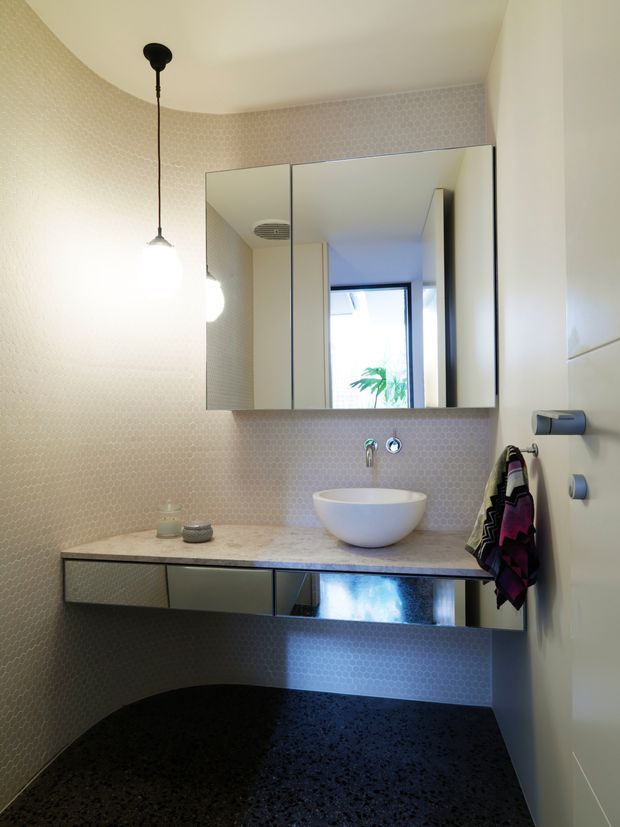 A compact contemporary bathroom with a floating vanity, white vessel basin, mirrored wall cabinet, pale small-format patterned wall tiles, dark speckled flooring, and a minimal pendant light. A window provides natural light, while a towel and wall-mounted fixtures are visible at the side.