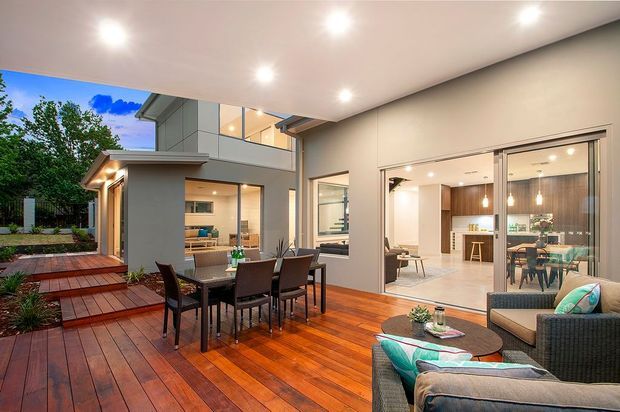 Contemporary two-storey home opening onto a spacious timber deck, with an outdoor dining setting and lounge furniture beneath a broad covered roof. Large glazed openings connect the deck to a bright open-plan interior featuring a kitchen, dining area and living space. Warm timber decking contrasts with light rendered walls, dark-framed glazing and landscaped planting.