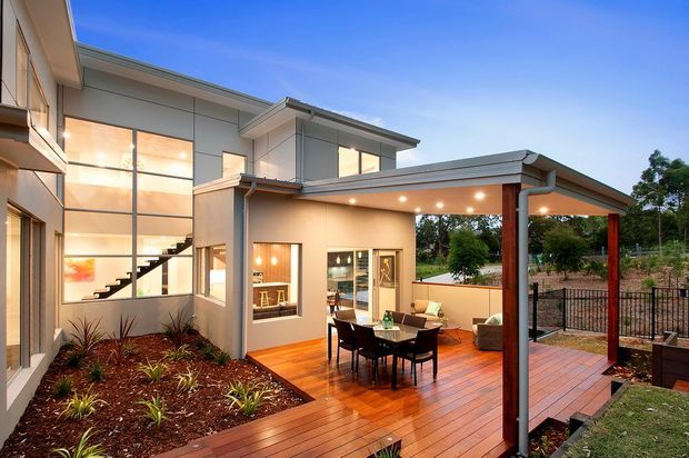 Contemporary multi-level home viewed from the landscaped rear garden, featuring large glazed walls, a projecting upper storey, and an expansive covered timber deck. The deck includes an outdoor dining setting and lounge area, with warm recessed lighting beneath the skillion-style roof overhang. A landscaped garden bed and fenced suburban outlook frame the composition.