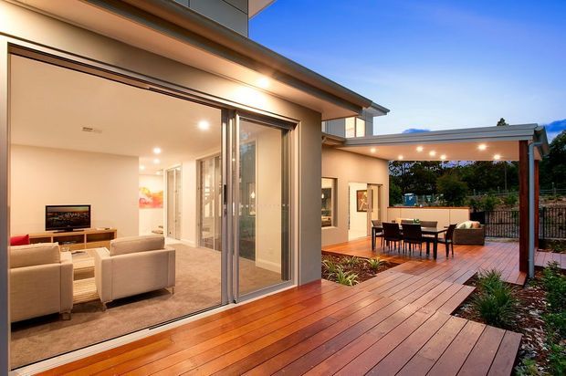 A contemporary home viewed from a timber deck at dusk, with large sliding glass doors opening to a bright living room. The covered outdoor entertaining area includes a dining table, lounge seating, recessed lighting and landscaped garden beds. Clean horizontal rooflines, dark-framed glazing and warm timber decking define the design.