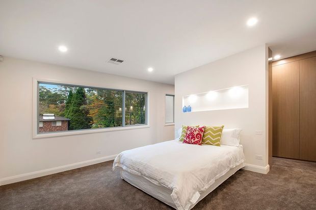 Bright contemporary bedroom with a low-profile upholstered bed dressed in white bedding, colourful patterned accent cushions, plush grey carpet, white walls, recessed ceiling lighting, a long horizontal window overlooking greenery, and tall built-in timber wardrobes. A recessed wall niche provides display shelving beside the bed.