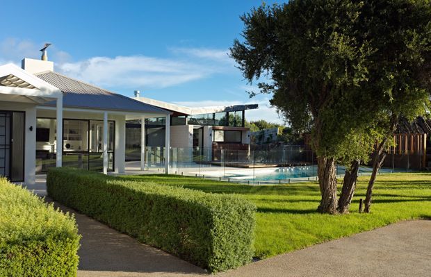 Exterior view of a contemporary residential property with a low-pitched corrugated roof, extensive glazing, a covered outdoor living area, manicured hedges and lawn, and a fenced swimming pool. The image is captured from the landscaped front or side garden, with the house and pool forming the main subject.
