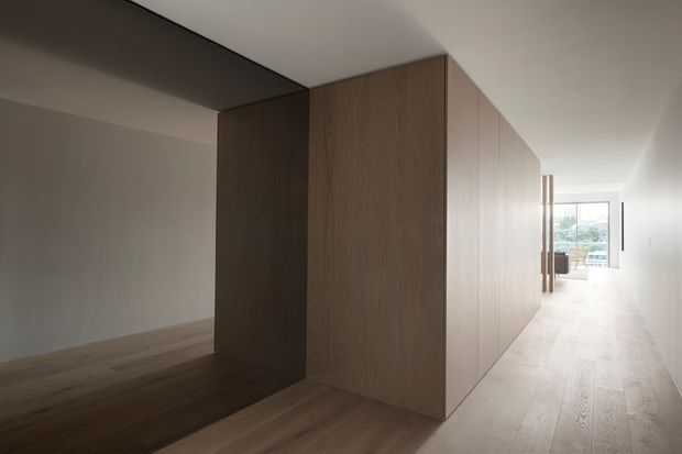 Minimalist interior corridor with a large continuous dark timber built-in volume dividing the space. Pale timber flooring extends through the hallway and adjacent open room, with white walls and ceiling and a bright glazed opening at the far end. The architectural composition is clean-lined and understated, with concealed or flush joinery and strong geometric planes.