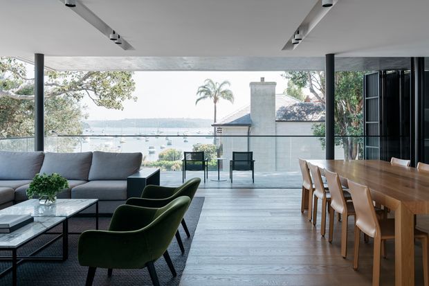 A sophisticated open-plan living and dining area with expansive floor-to-ceiling glazing overlooking a calm waterfront and boats. The room features a large grey sectional sofa, dark green lounge chairs, black-framed glazing and doors, a long timber dining table with timber chairs, pale timber flooring, and a streamlined white ceiling with recessed linear track lighting. A covered outdoor terrace and neighbouring roofline are visible beyond the glazing.