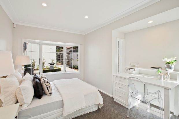Bright, refined bedroom with a white upholstered bed, layered neutral bedding and cushions, light walls with decorative ceiling cornice, plush grey carpet, and a built-in white desk and drawer unit positioned beneath a large window. French doors and glazing bring in abundant natural light, while a transparent acrylic chair and minimal accessories create an airy, polished look.