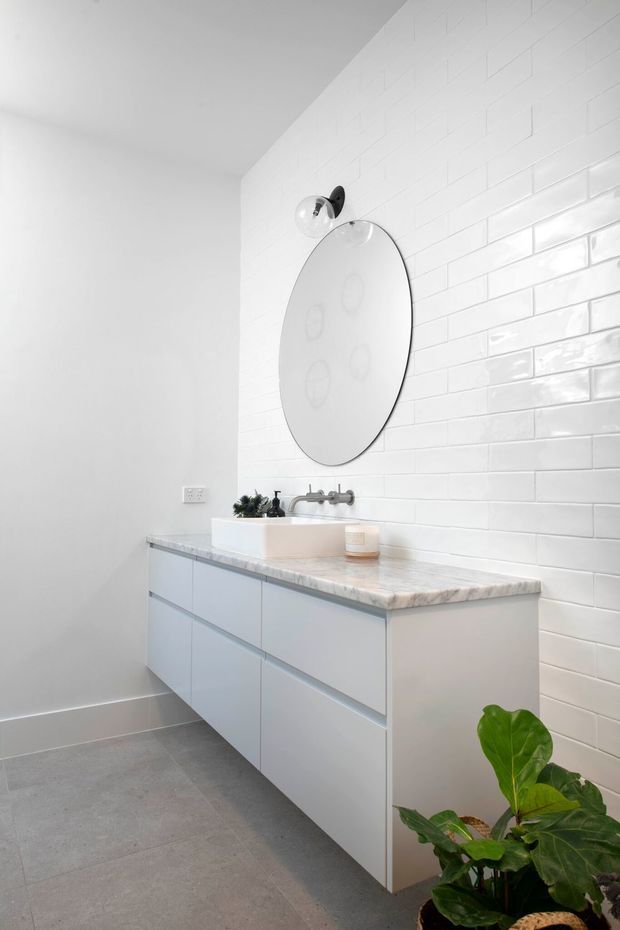 Bright contemporary bathroom vanity with a wall-mounted white floating cabinet, pale stone benchtop, integrated basin and chrome wall-mounted tapware. The feature wall is clad in glossy white subway tiles, complemented by a large oval mirror, white painted walls, and grey floor tiles. A potted plant adds greenery in the foreground.