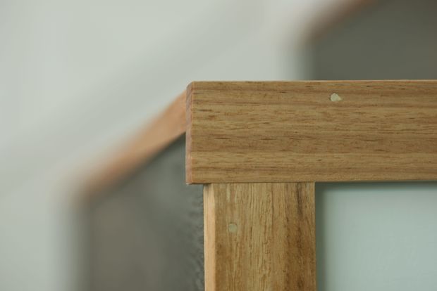 Close-up architectural detail of a warm-toned timber stair or balustrade, showing a horizontal handrail joined to a vertical timber post. The wood grain, exposed joinery, and softly blurred background dominate the image.